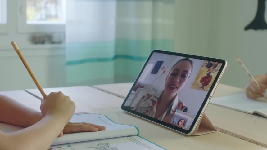 Two children at home do schoolwork while one watches their teacher on a tablet