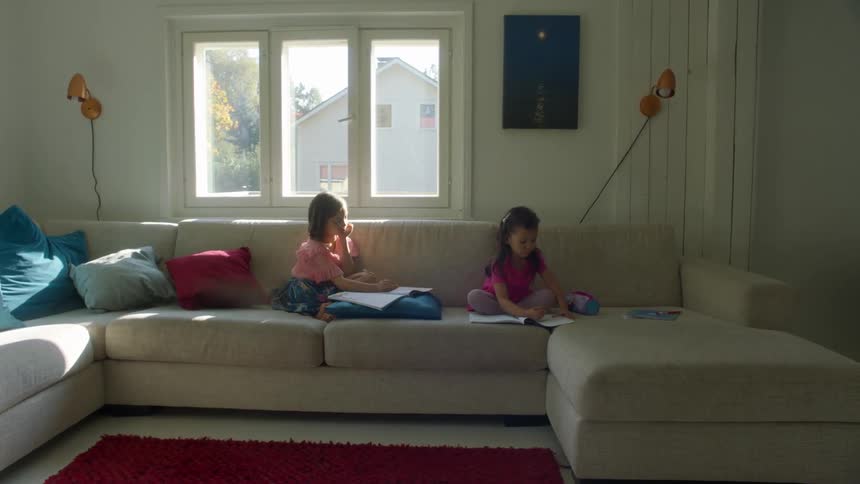 A pair of young girls write in softcover workbooks while sitting on a large sofa in a house during the day