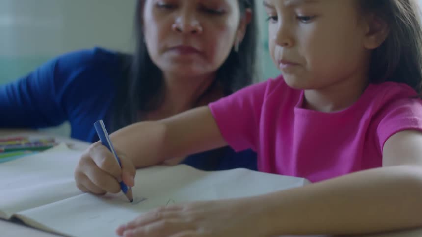 A young girl hands her mother a colored pencil while drawing and coloring at their kitchen table