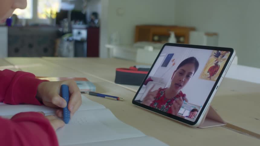 A child writes on paper sitting at a table at home while interacting with an adult on the screen of a laptop