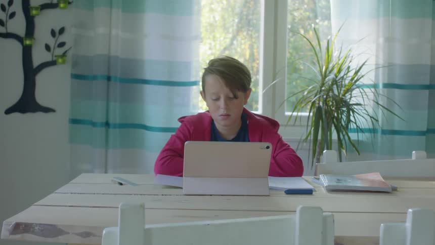 Boy with a red zip up hoodie sitting at table uses tablet and does homework in the living room at home