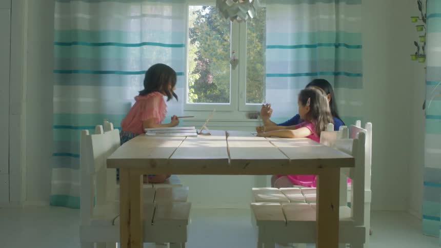 A woman kneels beside two young girls sitting at a table with pencils and books