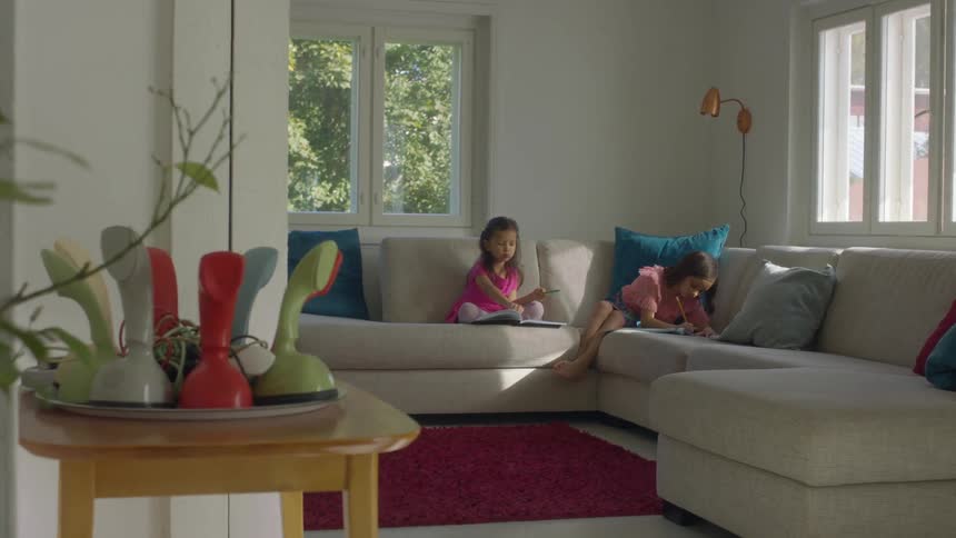 Two little girls sit on a couch at home and color in books