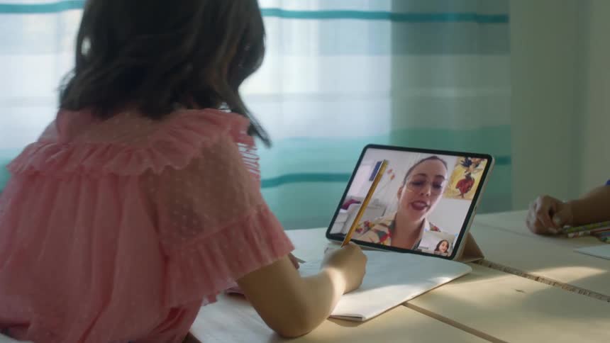 A young girl at home watches her teacher on a tablet as she does school work