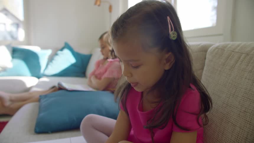 A girl in a house uses a colored pencil to color in a coloring book as she sits on a sofa next to her older sister in the living room of a house