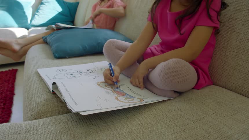 A young girl sitting crossed legged on a sofa uses a blue colored pencil to color an image in a coloring book