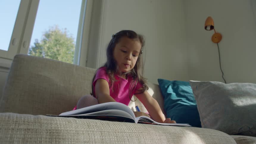 A young girl sits on the couch coloring or writing in a notebook