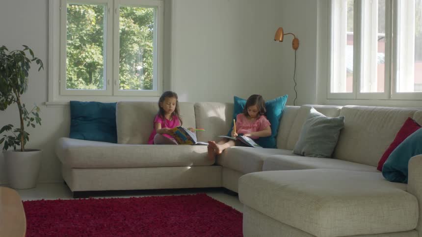 A pair of young girls write in workbooks while sitting on a large sofa in the living room of a house