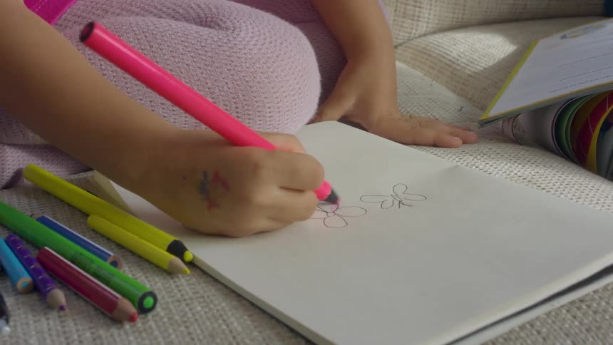 A small girl sitting on a sofa uses a pink colored pencil to color in a drawing on a sheet of paper