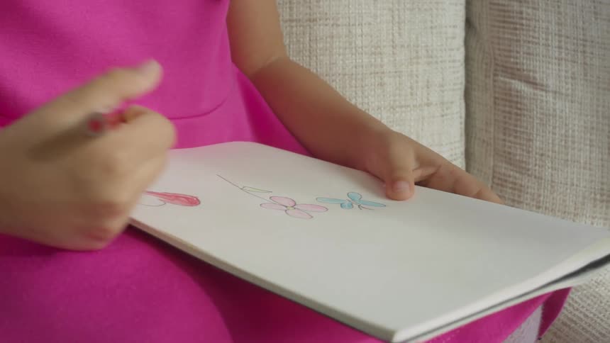 A girl in a hot pink dress drawing flowers in a notebook while sitting on a sofa
