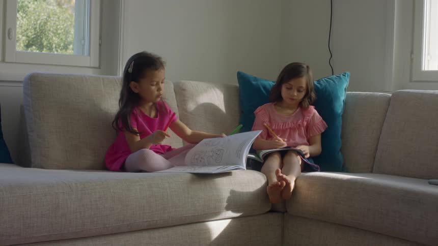 Two young girls sitting in a couch in the living room at home drawing in coloring books