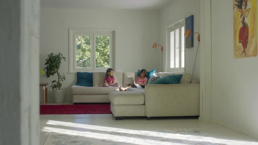 Two little girls sitting on a beige couch in a living room doing homework together