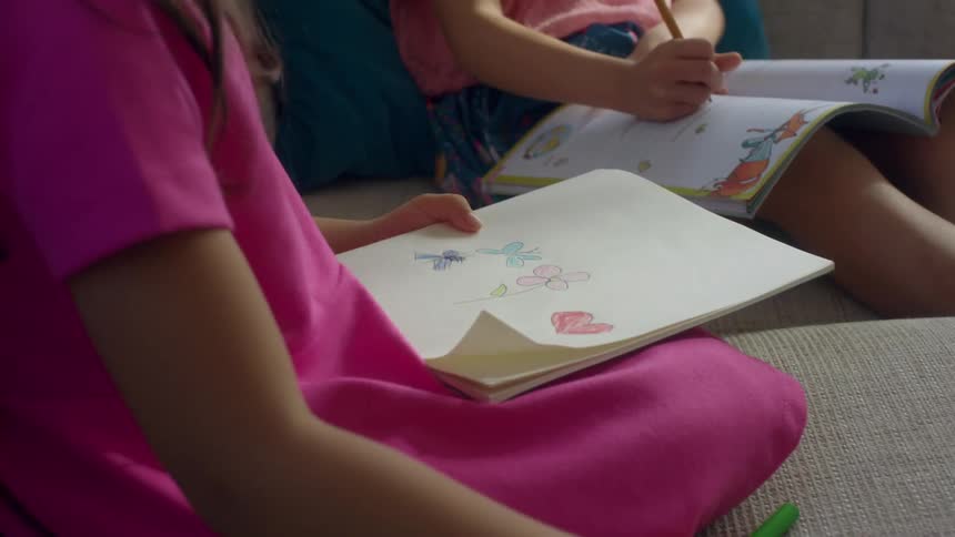 A young girl draws and colors while sitting on the couch next to a sibling doing homework
