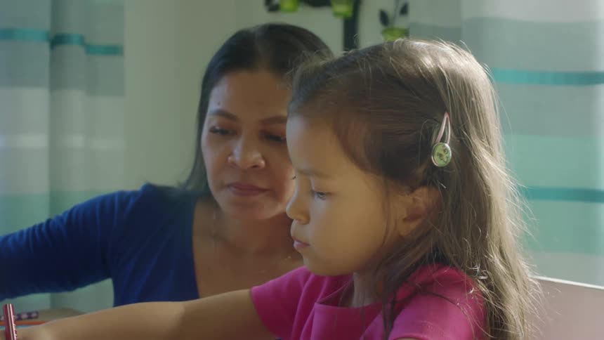 A mother sits next to her daughter at home and hands her a colored pencil while the little girl colors