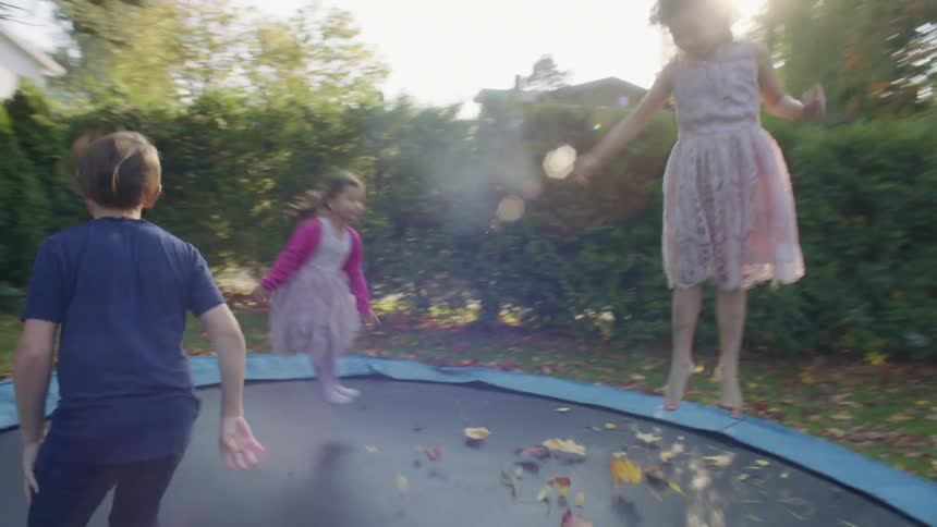 Parents watch children jump on trampoline in backyard bordered by hedge during day