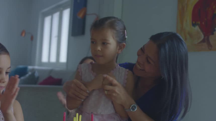 Candles on a birthday cake are blown out by a little girl with her mom and others watching