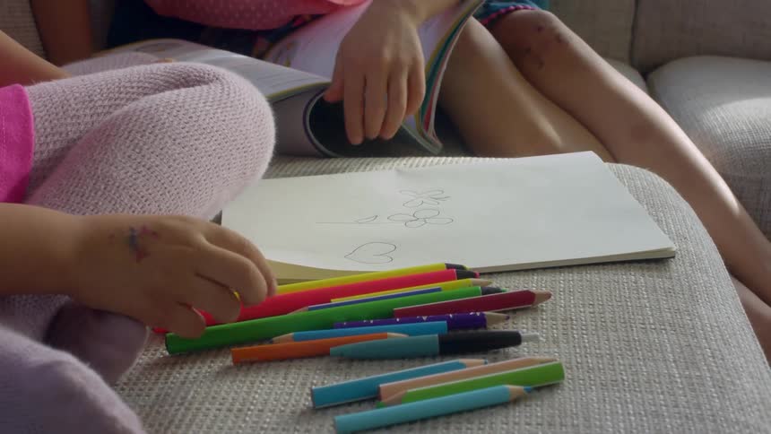 A child points to another childs drawing pad as they color while sitting on a couch indoors during the day