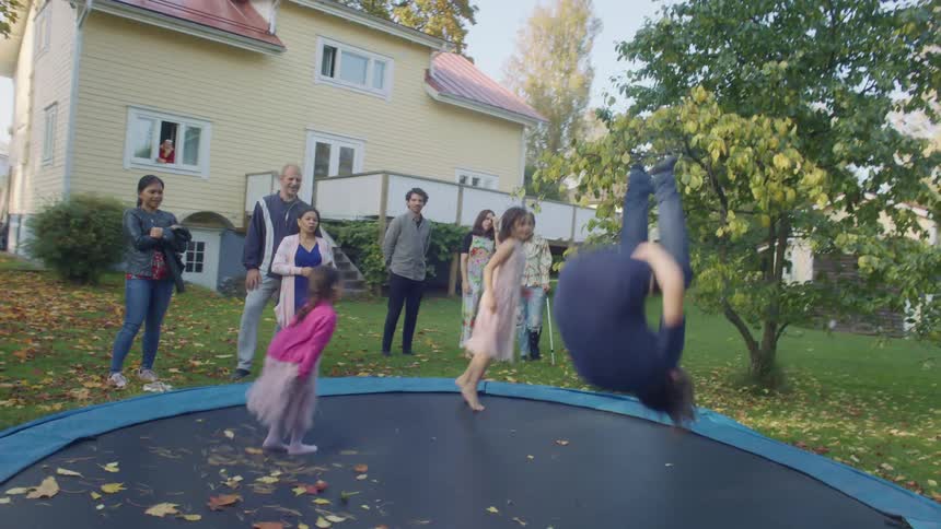 Three children jump on a trampoline in a back yard as their parents stand and watch
