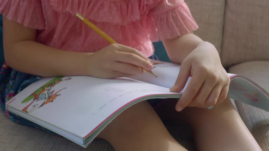 A young girl writes in an educational workbook with a pencil while sitting on a sofa