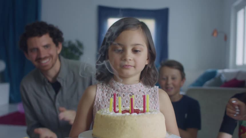 A young girl blows out the candles on a birthday cake with her family clapping behind her
