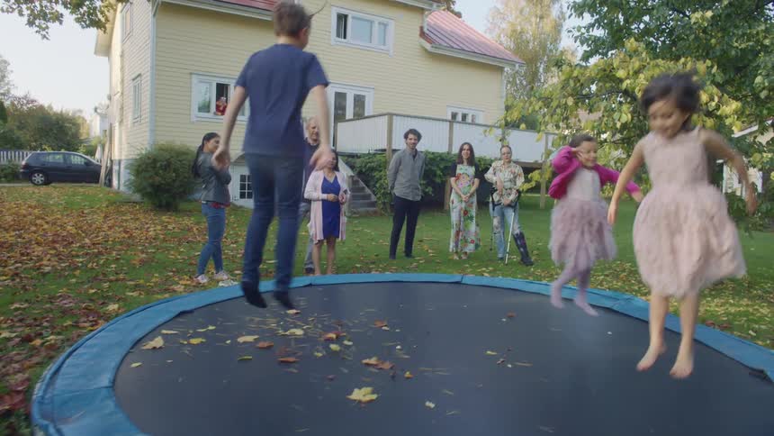 A group of kids jump on a trampoline in a large backyard while family watches lovingly