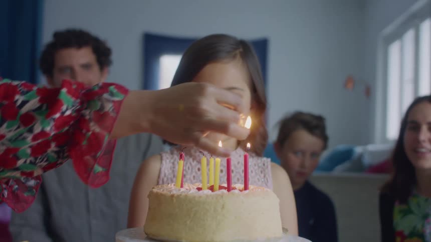 A young girl stands behind her birthday cake and smiles as her mother lights the candles