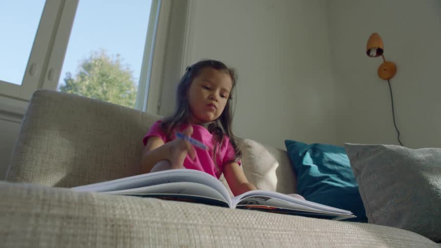 A young girl sits on a couch writing in a book in front of a window during the day