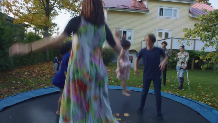 A group of children jump on a trampoline in the backyard of a house as adults stand nearby and watch