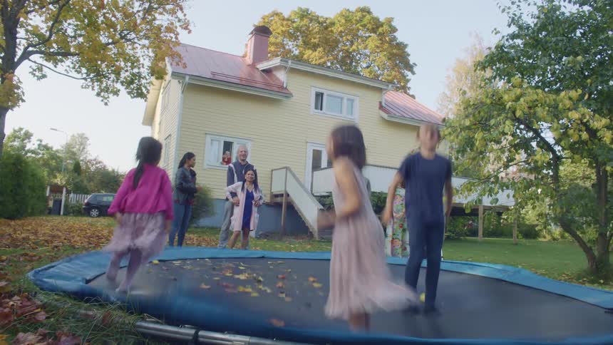 Two young girls and a boy jump on a trampoline in a backyard with multiple adults watching them