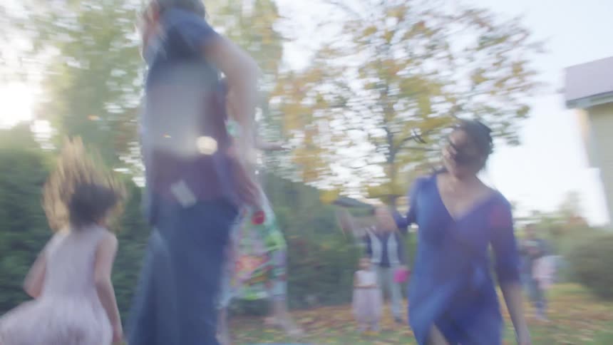 A group of children jump in a circle on a small trampoline in the backyard of house as adults watch from nearby during the day