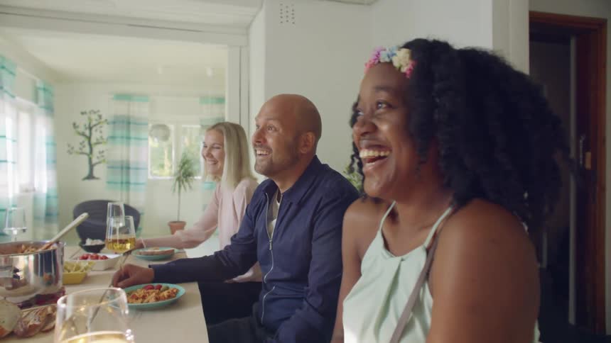 People laugh and talk as they sit at a table with food and wine in a home