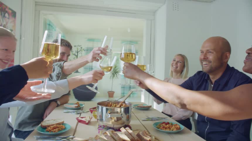 A group of people sit at a table with food on it and clink their wine glasses against one another