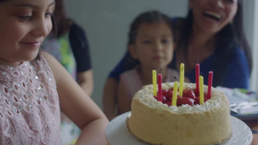 A girl blows out candles on a cake while kids and an adult clap and cheer indoors