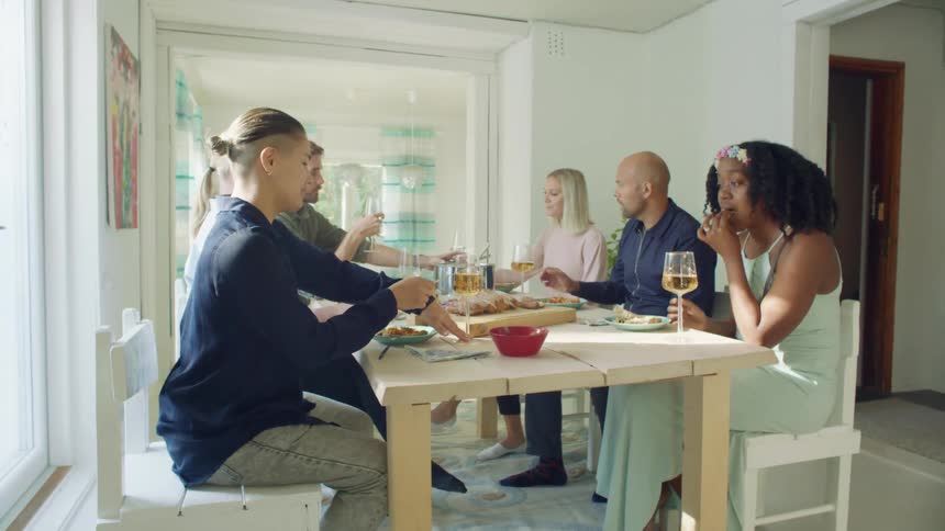 A group of people sit at a table in a home and eat and drink while talking