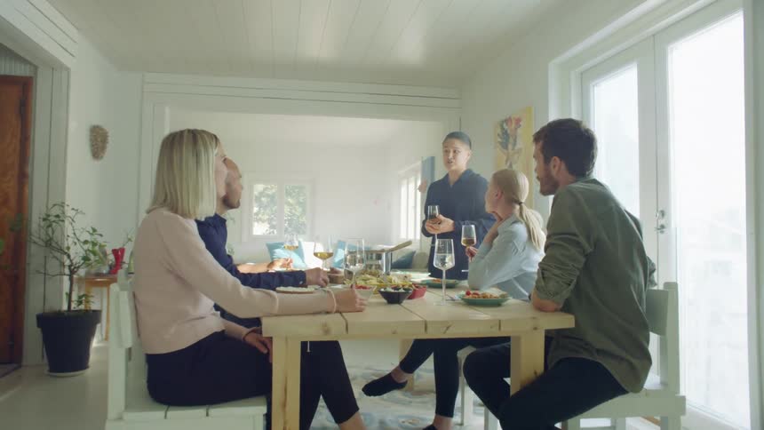 A woman holding a glass of wine stands at the head of a table in a house and talks to five seated people
