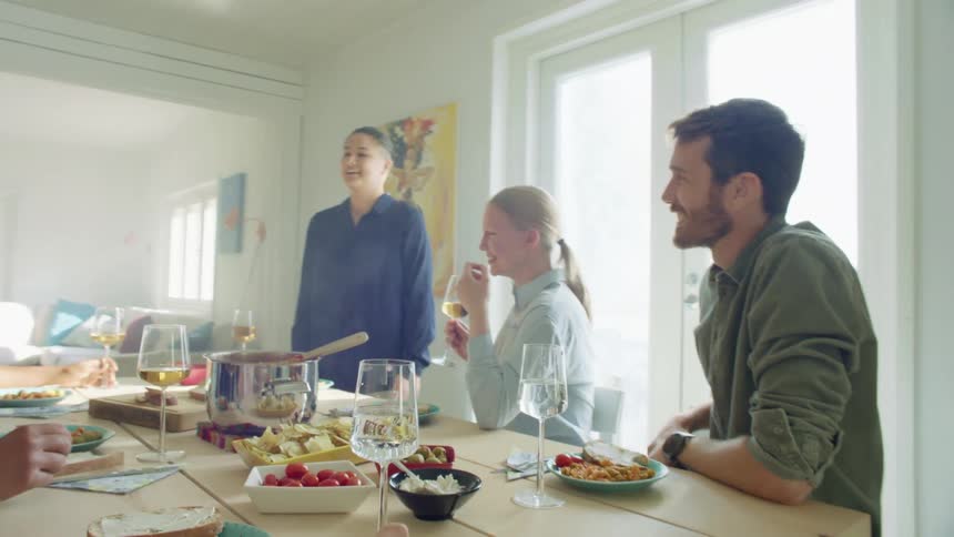 A young woman stands at a table and talks to a group of friends as they talk and drink during a meal