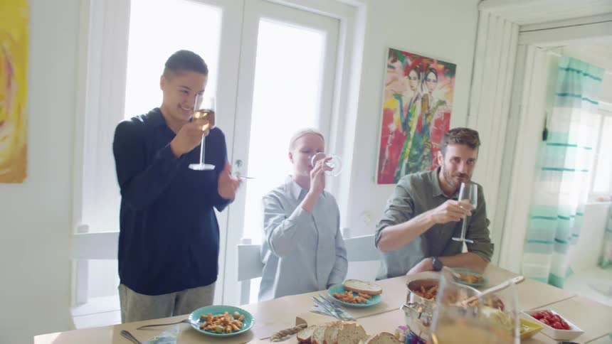 People sit or stand around a table covered with food in a bright sunlit room toast then drink