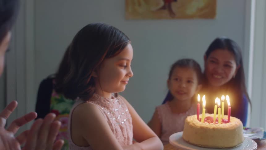 A group of people sing to a little girl as a birthday cake with lit candles is placed before her and she blows them out