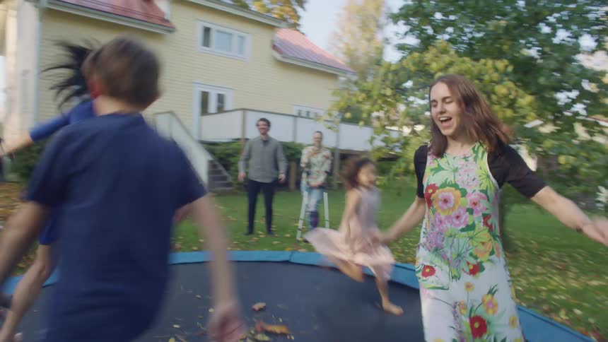 Kids run and play on a trampoline in a backyard as adults watch standing on the grass