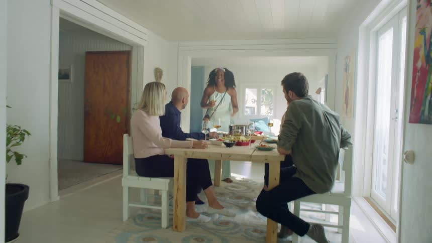A black woman stands at the end of a dining table talking to to others sitting at the table after eating