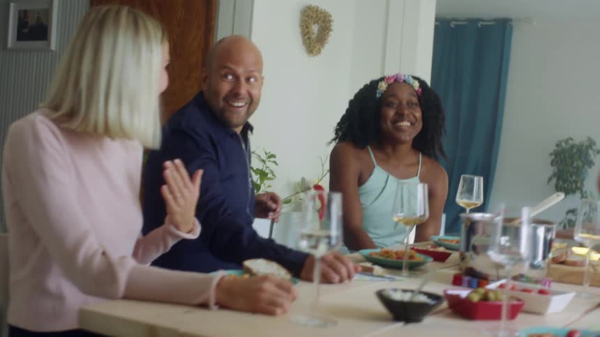 A group of friends laugh as they sit at a table drinking wine and eating