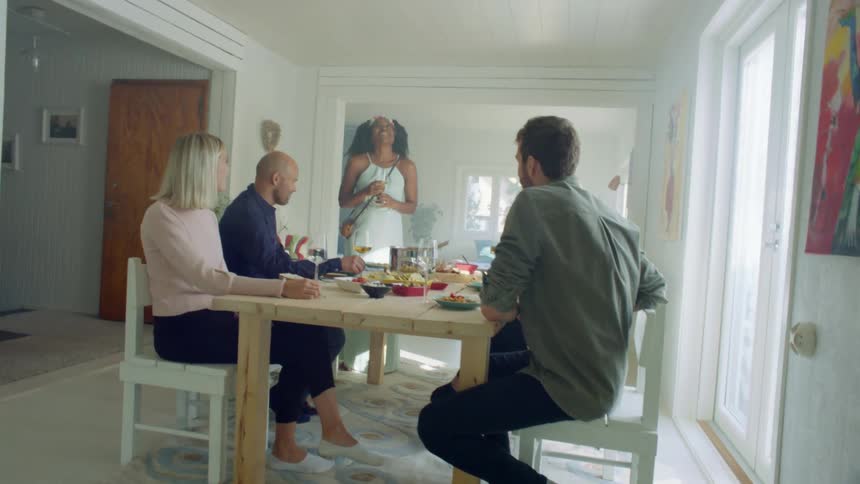 A woman standing in the dining room of a house talks to a group of people sitting around the dining table