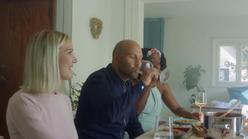 People sit at a table talking and laughing one man takes a drink of wine