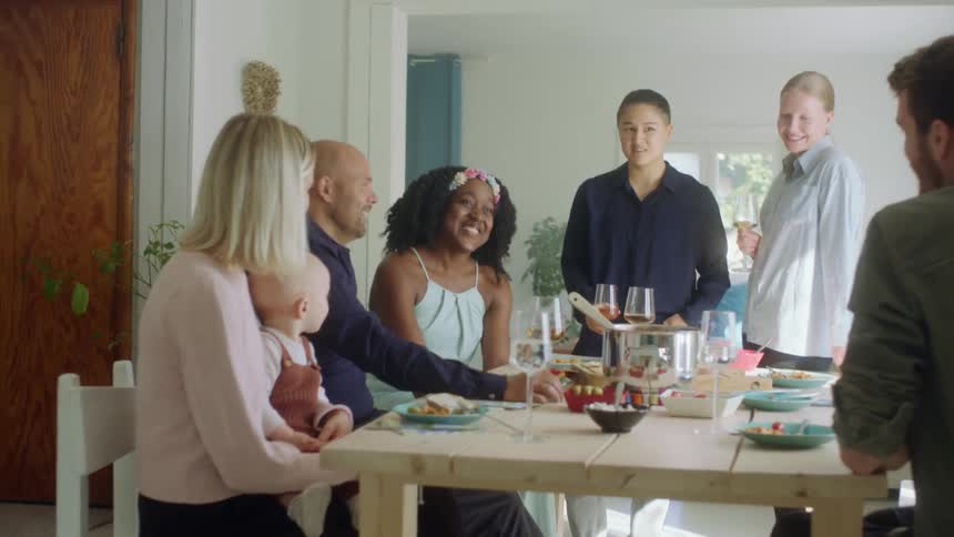 A group of people in a home sit around a table and talk and drink wine
