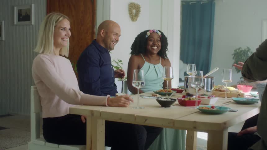 A group of men and women laugh and talk as they sit at a dining table at home enjoying dinner
