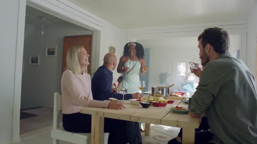 A group of people gather around a home dinner table with food and wine laughing and talking