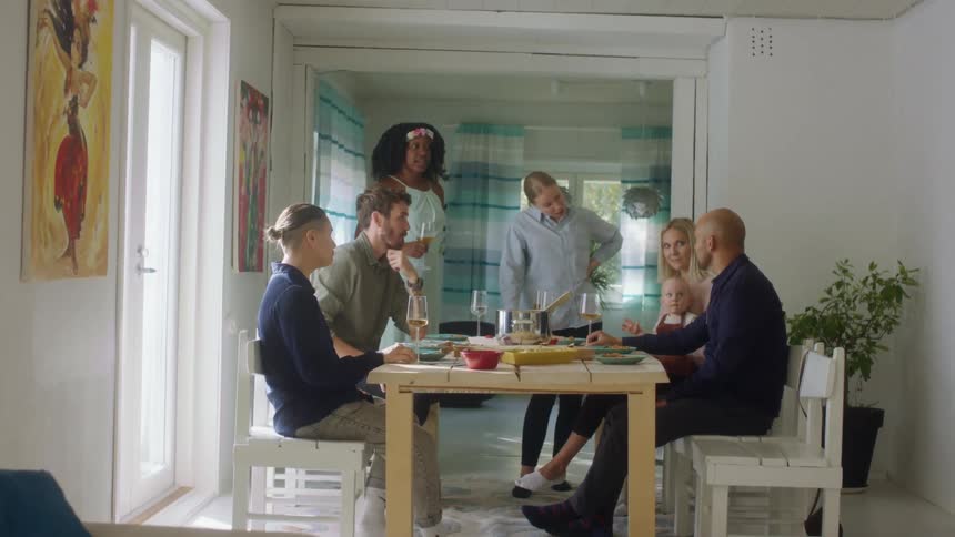 A group of people in a home gather around a table and eat and drink and talk