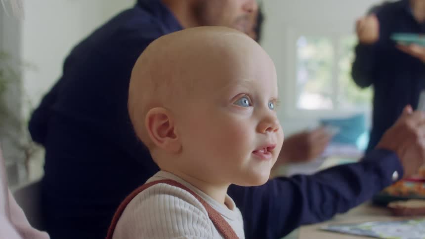 A group of people gather for food and wine at a home with a small baby happily smacking the table