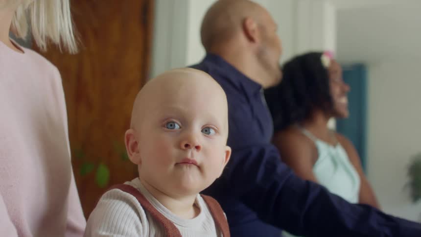 A baby sits on his mothers lap at a table with others as they talk and drink wine