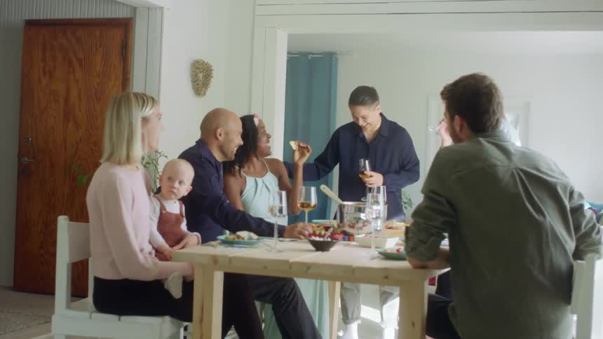 A group of people gathered around a table in a home eat and drink wine and talk